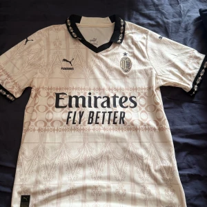 AC Milan x Puma beige fotbollströja M - AC Milan fotbollströja i beige med unikt grafiskt mönster och svarta detaljer på krage och ärmslut. Puma-logga, klubbmärke och sponsortryck på bröstet. Tillverkad i lätt och ventilerande funktionsmaterial, perfekt för match eller träning.