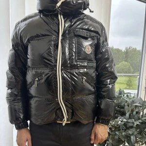 Svart Moncler pufferjacka med huva - Snygg svart pufferjacka från Moncler med glansig finish, stor huva och dragkedja framtill. Jackan har flera fickor med dragkedja och en bröstficka med Moncler-logga. Perfekt för kalla dagar när du vill hålla stilen.