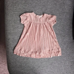 NY - Rosa plisserad klänning från KIWI - Supersöt ljusrosa plisserad klänning från KIWI med korta volangärmar och knappar i ryggen. Klänningen har glansigt tyg, rund hals och fina volangdetaljer längst ner. Perfekt för dig som gillar gulliga och trendiga plagg!