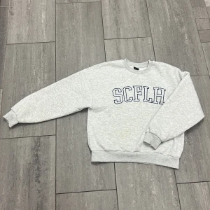 Grå sweatshirt SCFLH Gina Tricot M - Ljusgrå sweatshirt från Gina Tricot i storlek M med SCFLH-tryck i mörkblått framtill. Klassisk rund halsringning, ribbade muddar och mjuk insida. Perfekt för en chill och avslappnad look.