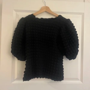 Svart puffärmig stickad tröja - Supersnygg svart tröja med puffiga trekvartsärmar och coolt stickat bubbelmönster. Tröjan har en lite boxig passform och är perfekt för dig som gillar statement-ärmar och unik struktur. Passar till både jeans och kjol för en trendig look.