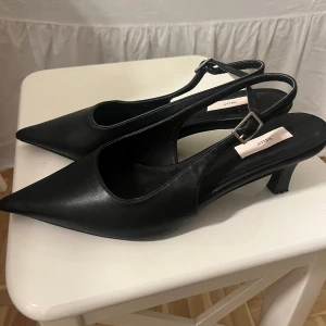 Svarta pumps med spetsig tå från Nelly - Stilrena svarta pumps från Nelly med spetsig tå och låg klack. Skorna har en öppen häl med justerbart spänne och är tillverkade i syntetmaterial med en elegant finish. Använda vid två tillfällen