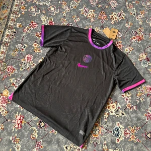 PSG x Nike svart fotbollströja L - Svart PSG x Nike fotbollströja i Dri-Fit-material med lila och rosa detaljer på krage och ärmslut. Bröstet har PSG-logga och en rosa Nike Swoosh med drip-effekt. Tröjan är kortärmad och har en sportig, modern look. Perfekt för dig som gillar unika matchtröjor.