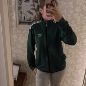 Grön Adidas track jacket med vita ränder - Säljer en mörkgrön Adidas track jacket med klassiska vita ränder på axlarna och dragkedja framtill. Jackan har hög krage, lång ärm och Adidas logga på bröstet. Perfekt för en sportig och chill stil.
