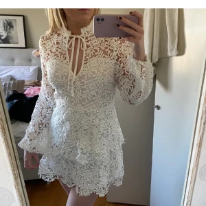 Vit klänning med volanger, studentklänning - Säljer en superfin vit spetsklänning med volanger och knytning vid halsen. Klänningen har långa ärmar och en kort längd, perfekt för studenten och sommarens fester. Aldrig använd.