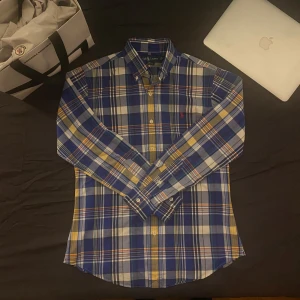 Blårutig skjorta från Ralph Lauren - Snygg blårutig skjorta från Ralph Lauren med klassisk krage och knappar framtill. Skjortan har långa ärmar och ett broderat rött märke på bröstet. Perfekt för en stilren look!