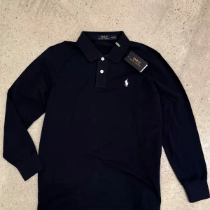 Mörkblå pikétröja från Ralph Lauren - Säljer en stilren mörkblå pikétröja från Ralph Lauren med klassisk krage och knappar framtill. Tröjan har långa ärmar och det ikoniska logotypbroderiet på bröstet. Perfekt för en avslappnad men ändå elegant look. Storlek M