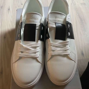 Valentino Garavani sneakers i vitt och svart - KVITTO FINS Snygga Valentino Garavani sneakers i vitt med en bred svart rem över snörningen. Skorna har en stilren design med vita snören och en diskret logga på plösen. Perfekta för en trendig look. 