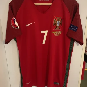 Röd Portugal fotbollströja från Nike Ronaldo - Säljer en röd Portugal fotbollströja från Nike med nummer 7 på framsidan. Tröjan har korta ärmar och är tillverkad i Dri-FIT material för optimal komfort. Den har detaljer som matchdatum och plats broderat på bröstet. Perfekt för fotbollsfans! Äkta med streckkod