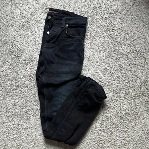 Nudie Jeans svarta - Snygga svarta jeans från Nudie Jeans med knappgylf och klassisk femficksdesign. Perfekta för en stilren look. Märkesdetaljer syns på bakfickan och i midjan. Storlek 29/34
