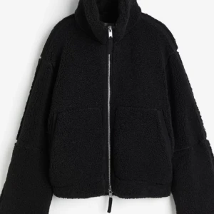 Svart teddyjacka med dragkedja - Mysig svart teddyjacka med dragkedja framtill och stora fickor. Perfekt för kyliga dagar med sin fluffiga och varma design. Jackan har en hög krage och långa ärmar för extra värme.