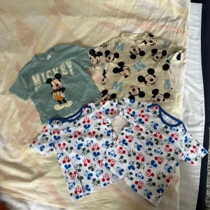 Fyra t-shirts med Musse Pigg-motiv - Säljer ett set med fyra t-shirts med Musse Pigg-motiv. En grön t-shirt med stor Musse Pigg-tryck och texten 'Mickey'. En beige t-shirt med flera Musse Pigg-ansikten och bokstaven 'M'. Två vita t-shirts med färgglada Musse Pigg-tryck i rött, blått och grått.