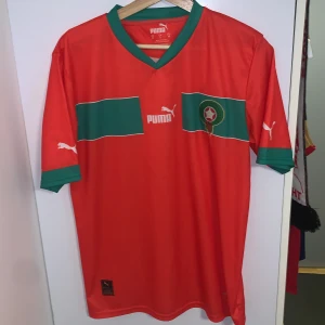 Röd fotbollströja från Puma Marocko  - Säljer en röd fotbollströja från Puma med gröna detaljer och korta ärmar. Tröjan har ett emblem på bröstet och Puma-loggor på ärmarna. Perfekt för fotbollsfans som vill visa sitt stöd. Marocko 