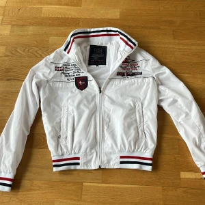 Vit jacka från Paul & Shark - Säljer en snygg vit jacka från Paul & Shark med broderade detaljer och dragkedja. Jackan har ribbade muddar och krage med röd och svart rand. Perfekt för sommaren. Nypris ligge på runt 5000 kr. Hör av er vid frågor eller funderingar 