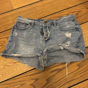 Blå jeansshorts med slitningar - Säljer ett par coola blå jeansshorts med slitningar och fransar. De har en högmidjad design och stängs med två knappar framtill. Perfekta för en avslappnad sommarlook!