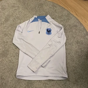 Frankrike tjocktröja från Nike - Säljer en vit långärmad tröja från Nike med ljusblå detaljer och dragkedja vid halsen. Perfekt för sportiga aktiviteter eller en avslappnad stil. Tröjan har en stilren design med ett emblem på bröstet. Köptes för 700kr. Pris kan diskuteras.
