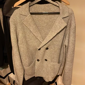 Grå stickad kofta från John Henric - Säljer en stilren grå stickad kofta från John Henric. Koftan har en dubbelknäppt design med två framfickor och ribbade detaljer. Perfekt för kyligare dagar men även skön att ha i varmare väder också och ger en elegant touch till din outfit.