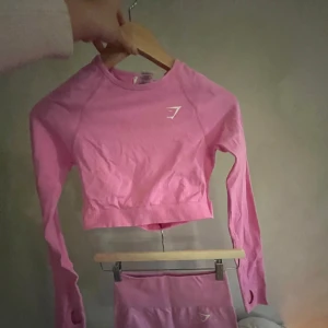 Rosa croppad träningströja och shorts från Gymshark - Säljer ett set med en rosa croppad träningströja och matchande shorts från Gymshark. Tröjan har långa ärmar och en stilren design med Gymshark-logga på bröstet. Perfekt för träning och att hålla stilen på gymmet! 💪