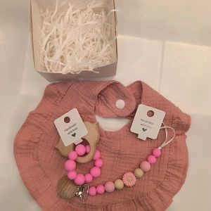 Napphållare present box - Charmigt handgjort set för barn med en rosa haklapp och matchande napphållare och bitring .Napphållaren har trä- och silikonpärlor i rosa och beige, samt en söt blomdetalj. Rosa haklapp är muslin med fin volang. Bitring är trä och silikon. Perfekt present i en fin förpackning.