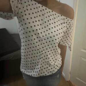 Vit offshoulder topp med svarta prickar - Säljer en söt vit offshoulder topp med svarta prickar. Toppen har korta ärmar och en elastisk kant som håller den på plats. Perfekt för en somrig look!