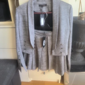 Grå kavaj och kjol från Supertrash - Snygg grå kavaj och matchande kjol från Supertrash. Kavajen har dekorativa knappar på ärmarna och en elegant krage. Kjolen har en stilren design som kompletterar kavajen perfekt. Perfekt för en stilfull look.