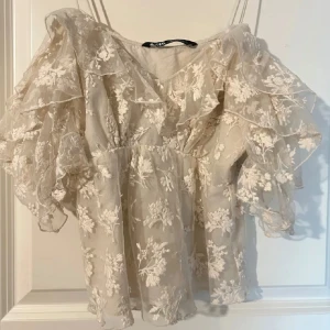 Beige blommig blus från Zara - Säljer en romantisk beige blus från Zara med vackra blommiga broderier. Blusen har volangdetaljer och en v-ringad design med trekvartsärmar. Perfekt för en elegant look.