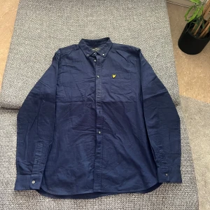 Mörkblå skjorta från Lyle & Scott - Snygg mörkblå skjorta från Lyle & Scott med klassisk krage och knappstängning. Skjortan har en broderad logotyp på bröstet och långa ärmar. Perfekt för en stilren look. Aldrig använd. Frakt tillkommer.