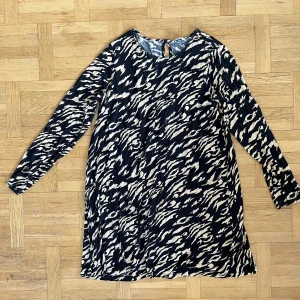 Mönstrad klänning från Indiska - Säljer en snygg mönstrad klänning från Indiska i storlek L. Klänningen har ett djärvt mönster i svart och beige med långa ärmar och en knappdetalj i nacken. Perfekt för en stilfull look! Finns hund och katt i hemmet.