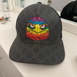 Gucci keps svart  - Snygg svart keps från Gucci med ett iögonfallande färgglatt örnmotiv på framsidan. Kepsen har ett diskret mönster och justerbart band baktill med röd och blå rand. Perfekt för att ge din outfit en unik touch. Dessutom så är kepsen limiterad vilket väldigt få människor har den och är sällsynt att få tag i. Har haft den i 3 år och den är i väldigt bra skick och självklart är den äkte. OBS den har ej kvitto eller låda.