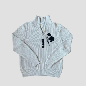 Beige Half Zip With Spades - Snygg beige stickad tröja med dragkedja och svart blomdetalj på bröstet. Perfekt för en stilren look med långa ärmar och ribbad struktur. Passar både herr och dam.