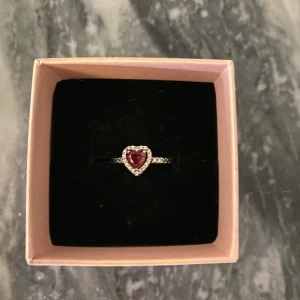 Ring med hjärtformad röd sten från Pandora - Elegant ring från Pandora med en hjärtformad röd sten i mitten, omgiven av gnistrande stenar. Perfekt för att ge en romantisk touch till din stil. Kommer i en fin presentask.