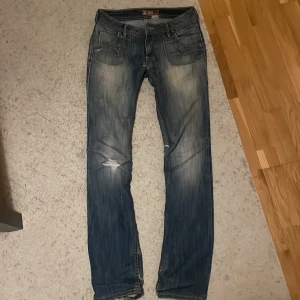 Blå jeansbyxor med slitningar - Snygga blå jeansbyxor med en cool slitning på ena benet. De har en klassisk femficksdesign och knappar med unika detaljer. Perfekta för en avslappnad stil. De har hål i mitten (visad på bild) men det går lätt att sy. De är dessutom sydda för att vara lågmidjade.