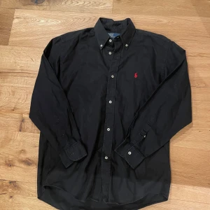 Svart skjorta från Ralph Lauren - Snygg svart skjorta från Ralph Lauren i 100% bomull. Skjortan har långa ärmar och knappar framtill. Den är dekorerad med ett rött broderat märke på bröstet. Perfekt för en stilren look.