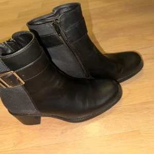 Svarta boots med dragkedja och spänne - Snygga svarta boots i skinn med dragkedja på sidan och ett dekorativt spänne. Perfekta för höst och vinter med en stilren design och bekväm passform. Aldrig använt helt nya!!