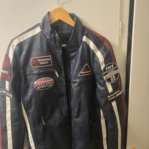 Svart läderjacka skinnimitation jacka med racingdetaljer mc - Säljer en svart skinnimitation jacka coola racingdetaljer och broderade märken. Jackan har en dragkedja framtill och långa ärmar. Perfekt för dig som gillar motorcykelstil! Fint skick. Stl S, normal I storleken. Vaddering på axlar och armbågar.  Det finns ytterligare en liknande läderjacka bland mina andra annonser, helt svart. 