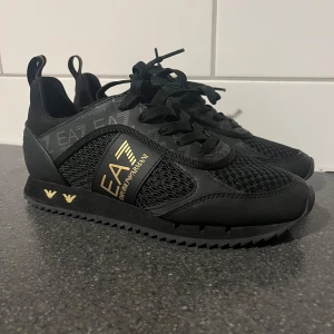 Svarta sneakers från Emporio Armani - Säljer dessa snygga skor från Armani med guld detaljer. Skorna är endast använda 2 gånger och nypris är 2100kr. Finns inga tecken på användning förutom på baksidan men det går nog att tvätta bort.