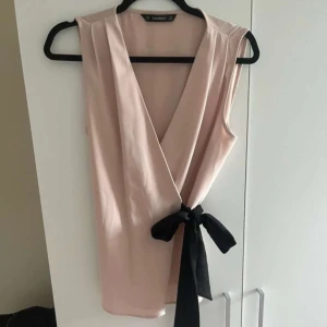 Beige omlottopp från Zara Basic - Snygg beige omlottopp från Zara Basic med en svart knytning i midjan. Toppen är ärmlös och har en djup v-ringning som ger en elegant touch. Perfekt för att klä upp eller ner beroende på tillfälle.