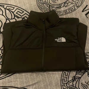 Olivgrön kofta från The North Face - Säljer en olivgrön kofta från The North Face med dragkedja och hög krage. Koftan har en praktisk ficka med dragkedja på ärmen och logotypen på bröstet. Perfekt för kyliga dagar.