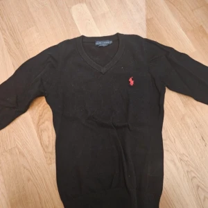Svart tröja från Ralph Lauren - Snygg svart tröja från Ralph Lauren med röd broderad logga på bröstet. Tröjan har en v-ringad hals och långa ärmar. Perfekt för en stilren look.