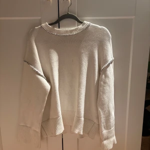 Vit stickad tröja med detaljer - Säljer en vit stickad tröja med unika svarta sömdetaljer från Dagmar. Tröjan har en avslappnad passform med långa ärmar och ribbade kanter vid hals, ärmslut och nederkant. Den är lite små smutsig, men ska försöka få bort fläckarna!🥰