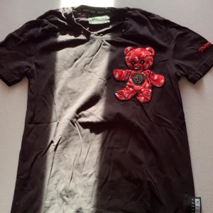 Svart Philip plein tshirt  - Cool svart t-shirt från Philipp Plein med ett stort rött nallemotiv på framsidan. T-shirten har korta ärmar och en rund halsringning. Perfekt för en unik och trendig stil.