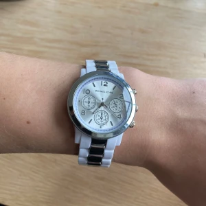 Klocka från Michael Kors - Snygg vit klocka från Michael Kors med silverdetaljer och tre mindre urtavlor. Armbandet är i rostfritt stål.  Perfekt accessoar för att lyfta din stil. Vet inte exakt hur mycket den kostar som nypris då jag har fått den. Men när jag söker upp på liknade klockor från samma märke kostar dom runt 1200kr. 