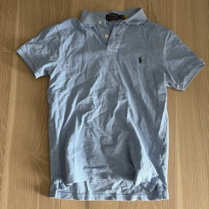 Ljusblå pikétröja från Ralph Lauren - Säljer en stilren ljusblå pikétröja från Ralph Lauren. Tröjan har en klassisk krage med två knappar och det ikoniska broderade logotypen på bröstet. Perfekt för en avslappnad men ändå snygg look.