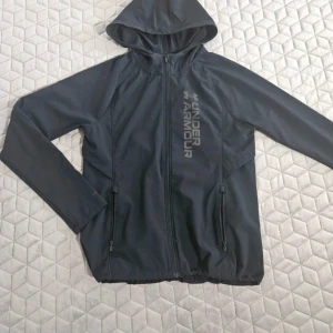 Under Armour Jacka  - Säljer en svart vindjacka från Under Armour med huva och dragkedja. Jackan har långa ärmar och är perfekt för löpning eller andra utomhusaktiviteter. Den är tillverkad i ett lätt syntetmaterial och har en stilren design med logotypen tryckt på framsidan. Modellen säljs inte längre så passa på 