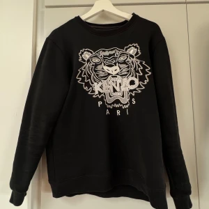 Svart sweatshirt från Kenzo med tigerbroderi - Säljer en svart sweatshirt från Kenzo med ett stort, detaljerat tigerbroderi och texten 'Kenzo Paris' på framsidan. Tröjan har långa ärmar och en rund halsringning. Perfekt för en stilren och trendig look.