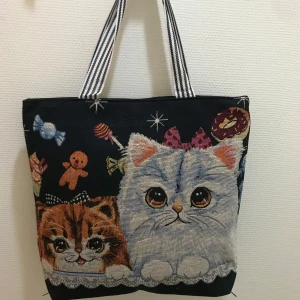 Canvasväska  med kattmotiv - Charmig väska med ett sött kattmotiv i olika färger. Väskan har randiga handtag och ett lekfullt mönster med katter, godis och stjärnor. Perfekt för kattälskare som vill ha en unik och rolig accessoar.  Helt ny 