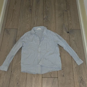 Randig skjorta från H&M - Säljer en stilren blå- och vitrandig skjorta från H&M. Skjortan har långa ärmar och knappar framtill. Perfekt för en avslappnad men ändå snygg look. 