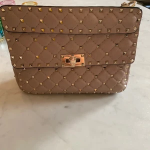 Valentino väska - Säljer nu min superfina väska i färgen poudre från Valentino med nitar. Väskan har en elegant kedjerem och ett lås framtill. Insidan är röd med en dragkedjeficka för extra förvaring. Nypris cirka 32 000 kr. Har dustbag!