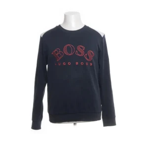 Svart sweatshirt från Hugo Boss - Snygg svart sweatshirt från Hugo Boss med röd text på bröstet. Tröjan har långa ärmar och en klassisk rund halsringning. Perfekt för en stilren och avslappnad look.