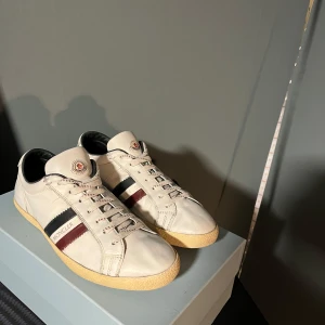 Moncler skor - Snygga vita sneakers från Moncler med klassiska ränder i svart och röd på sidan. Skorna har en stilren design med snörning och en bekväm sula. Perfekta för en casual look. Skulle säga att dom är i väldigt bra skick men man bedömer skick själv, självklart äkta men tyvärr inget og, skriv vid minsta fundering🌟